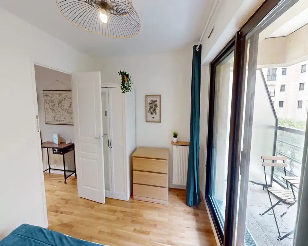 Bright, modern bedroom with wooden floors and a sliding door opening to a small balcony. Simple wardrobe and drawer unit, teal curtains and small plants create a calm, cozy atmosphere ideal for relaxing.