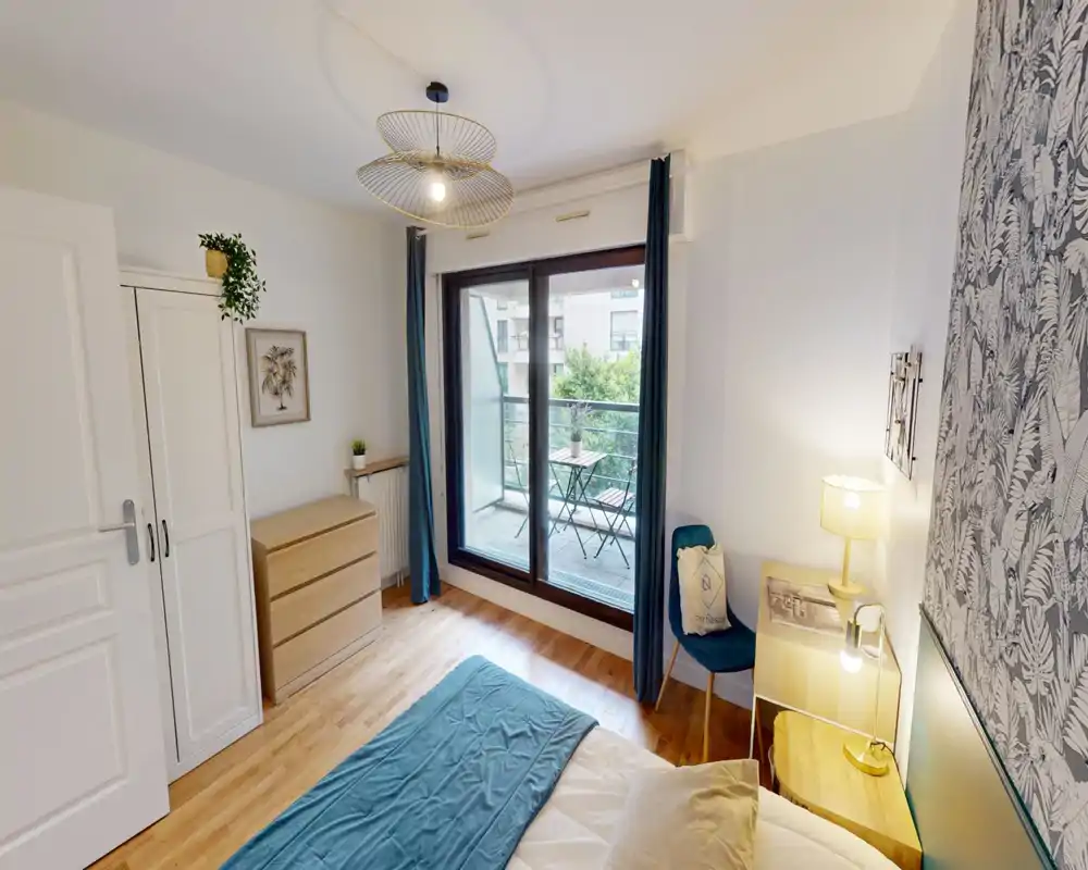 Bright, stylish bedroom with a comfortable double bed, bedside table and lamp, wardrobe and chest of drawers. Large sliding doors open to a small balcony with seating and greenery, letting in abundant natural light.