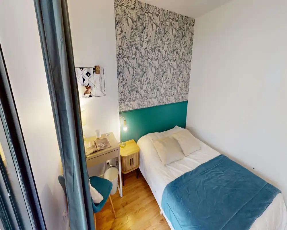 Bright, styled bedroom with a double bed, teal headboard and patterned wallpaper, bedside table and a small desk — cozy and well-lit for a short-term rental.