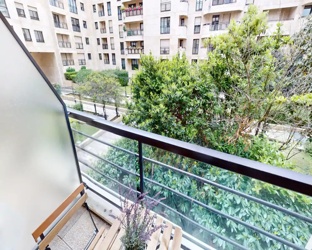 Sunny small balcony with metal railing, folding wood chairs and potted lavender, overlooking a landscaped courtyard and apartment facades.