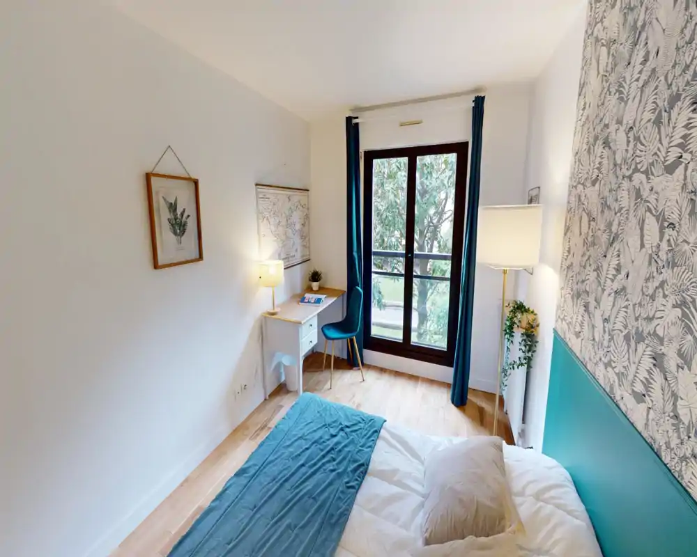 Chambre lumineuse et chaleureuse avec accent turquoise, papier peint décoratif et un petit bureau près de la fenêtre. La grande fenêtre apporte une belle lumière naturelle et une vue sur les arbres.