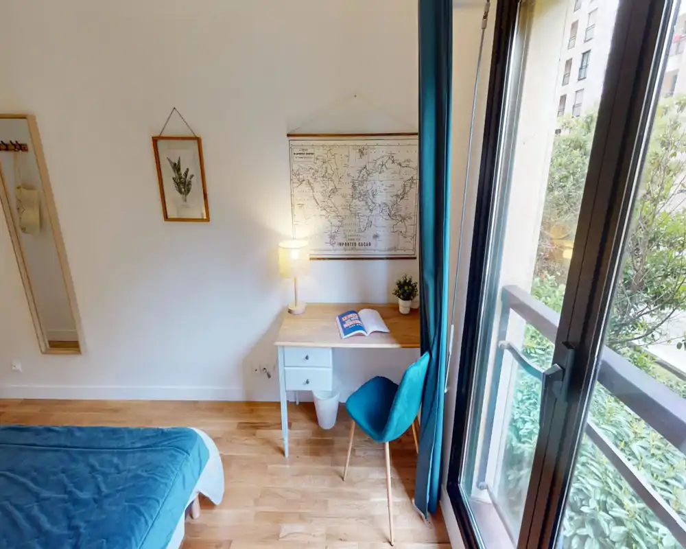 Coin chambre lumineux et chaleureux avec une couette bleu-vert, un petit bureau en bois et une grande fenêtre laissant entrer la lumière naturelle — idéal pour se reposer et travailler.