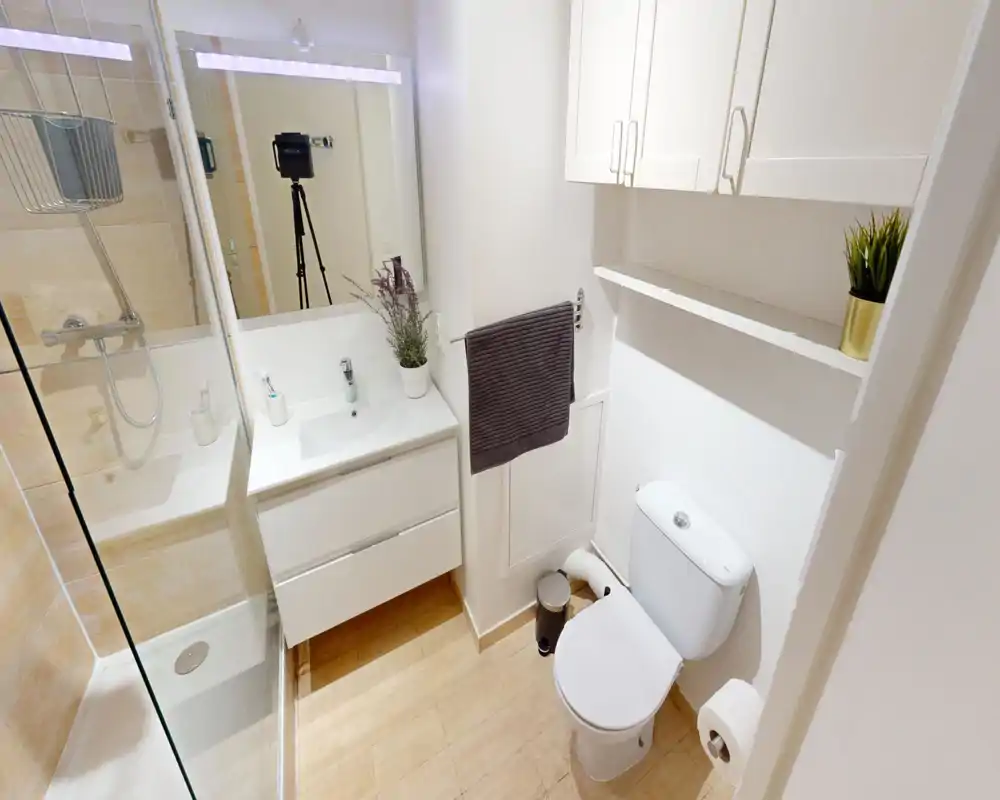 Salle de bain moderne et propre avec douche vitrée, meuble-lavabo blanc et WC ; carrelage clair et lumière vive pour une atmosphère aérée.