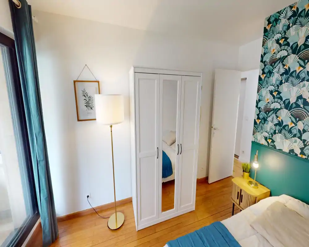 Bright, styled bedroom with a double bed, bedside table and decorative wallpaper. Large window provides natural light and a mirrored wardrobe adds depth.