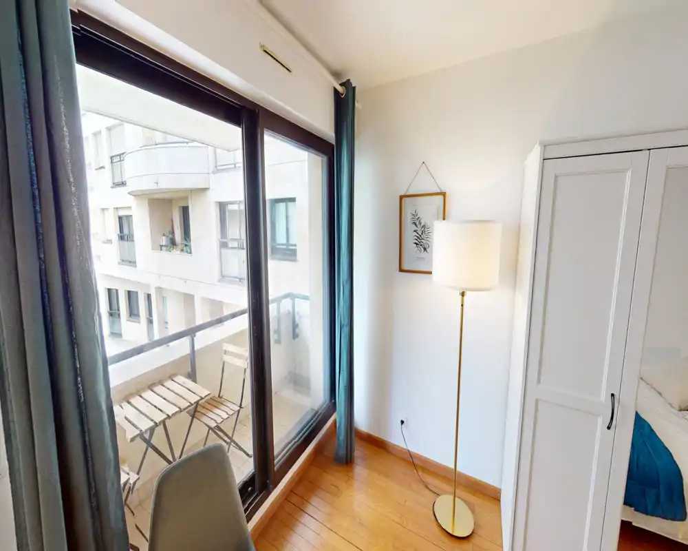 Bright bedroom corner with floor lamp, wardrobe and a glimpse of the bed next to large sliding windows opening onto a small balcony with bistro furniture.