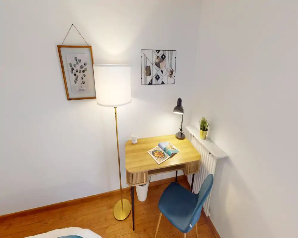 Small bedroom corner with a wooden desk and bedside area: a blue chair, warm floor and desk lamps, decorative wall art and a glimpse of the bed at the bottom of the frame.
