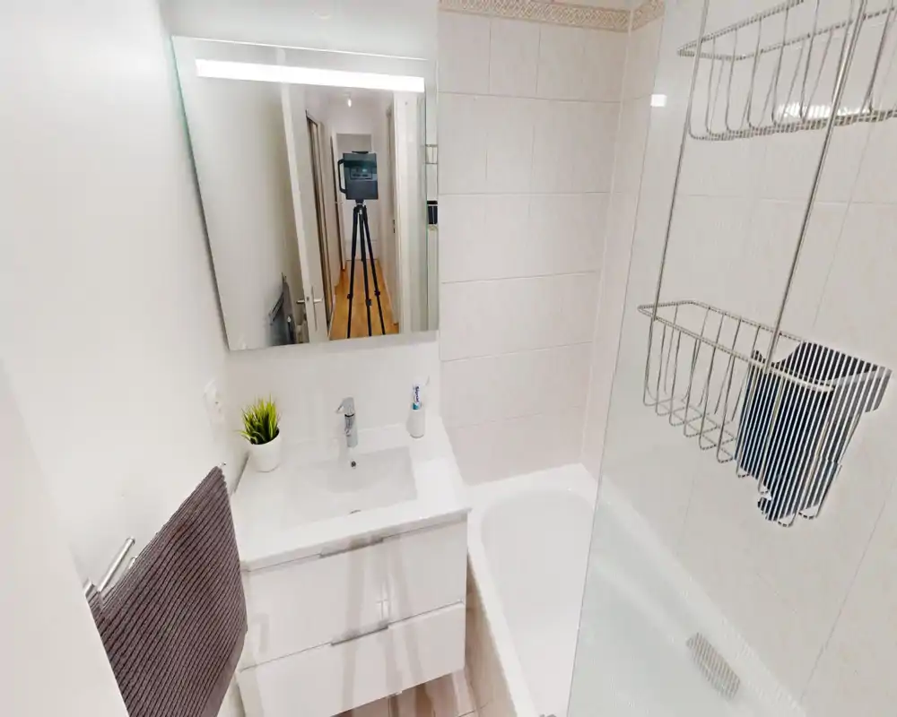 Bright, compact bathroom with a white sink vanity, mirror and bathtub with a glass shower screen. Clean modern tiles and practical storage make it suited for short-term rentals.