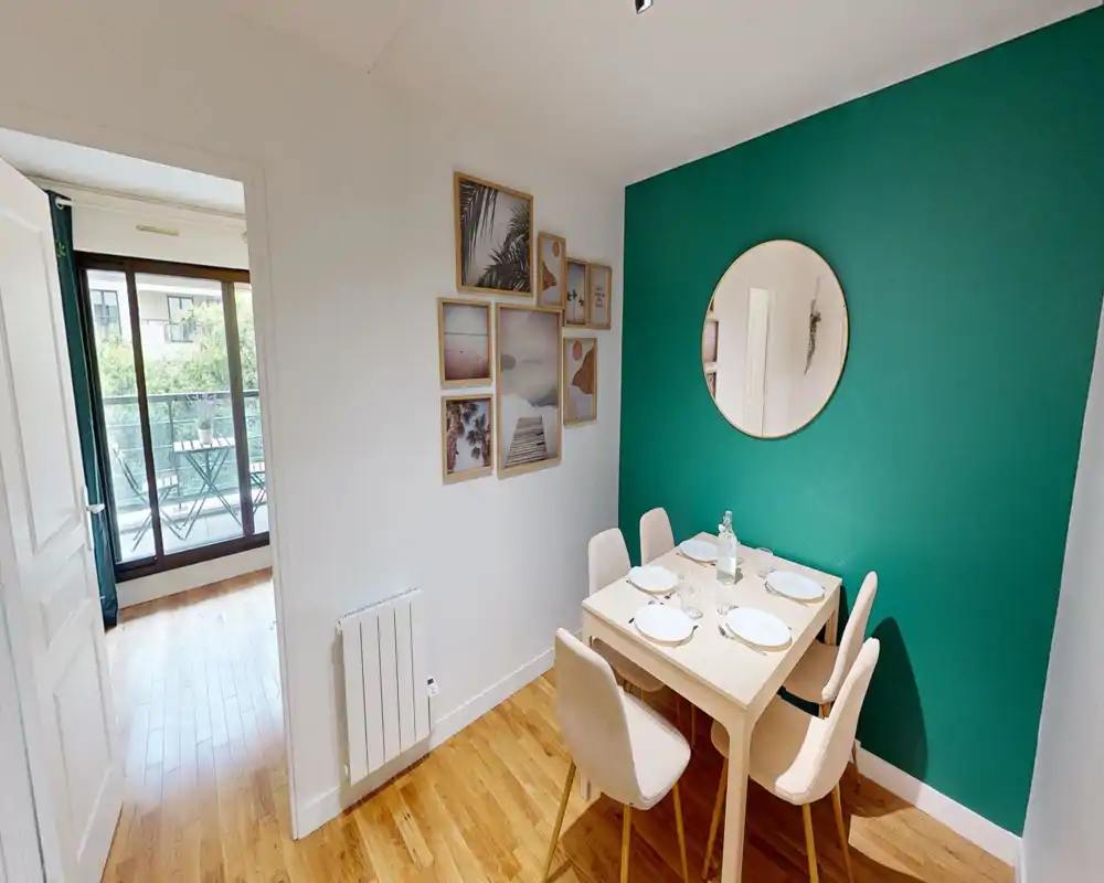 Cozy, well-staged dining corner with a small table for four, upholstered chairs, decorative wall art and a round mirror set against a teal accent wall — bright and welcoming for meals.
