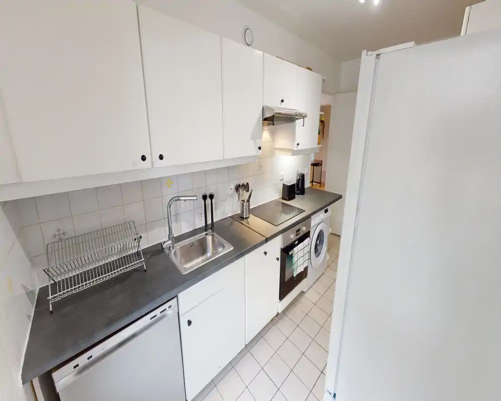 Bright, compact kitchen with white cabinets, a stainless sink, built-in oven and washing machine — practical and tidy for a small apartment.