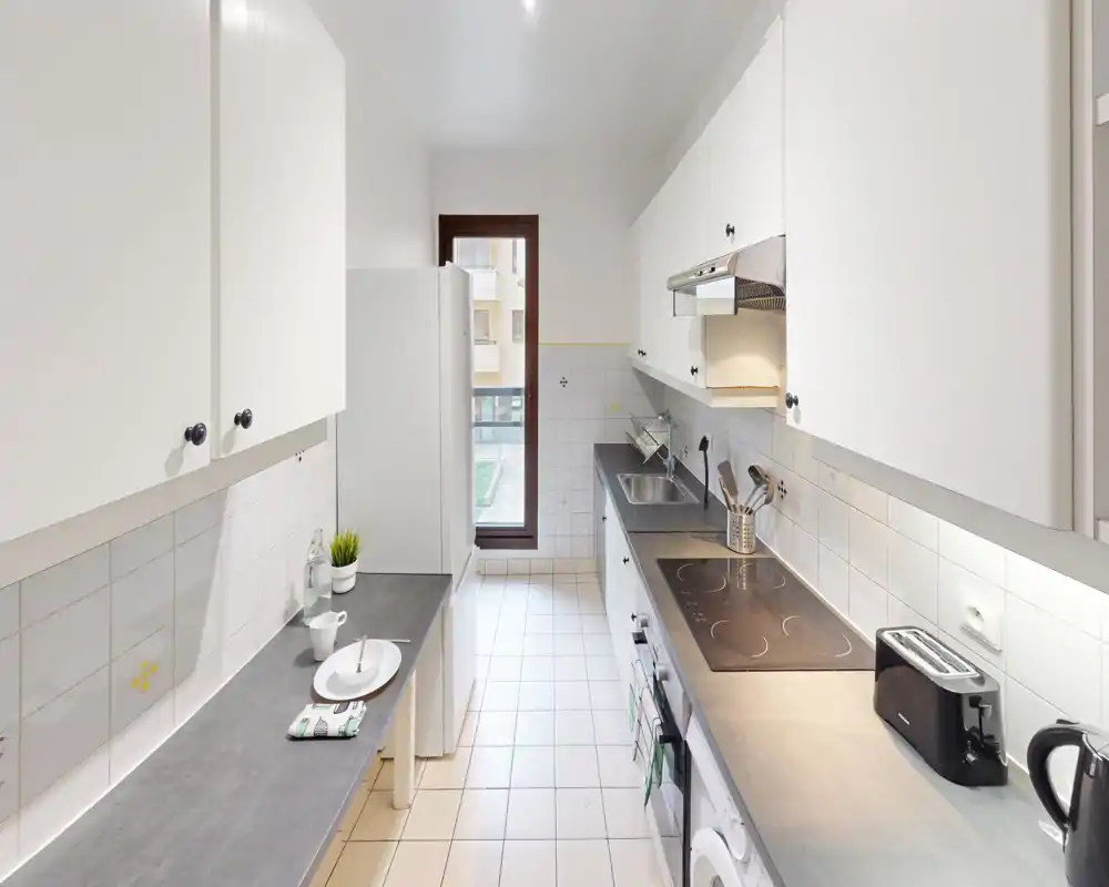 Bright, modern galley kitchen with white cabinets, integrated stovetop and a window at the far end. Clean countertops, toaster and kettle visible — ideal for rental listings highlighting practicality and neat styling.