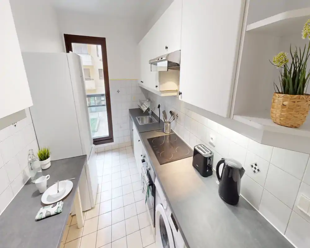 Bright, narrow galley kitchen with white cabinets, electric cooktop, fridge and small breakfast counter; modern and tidy layout ideal for a rental.