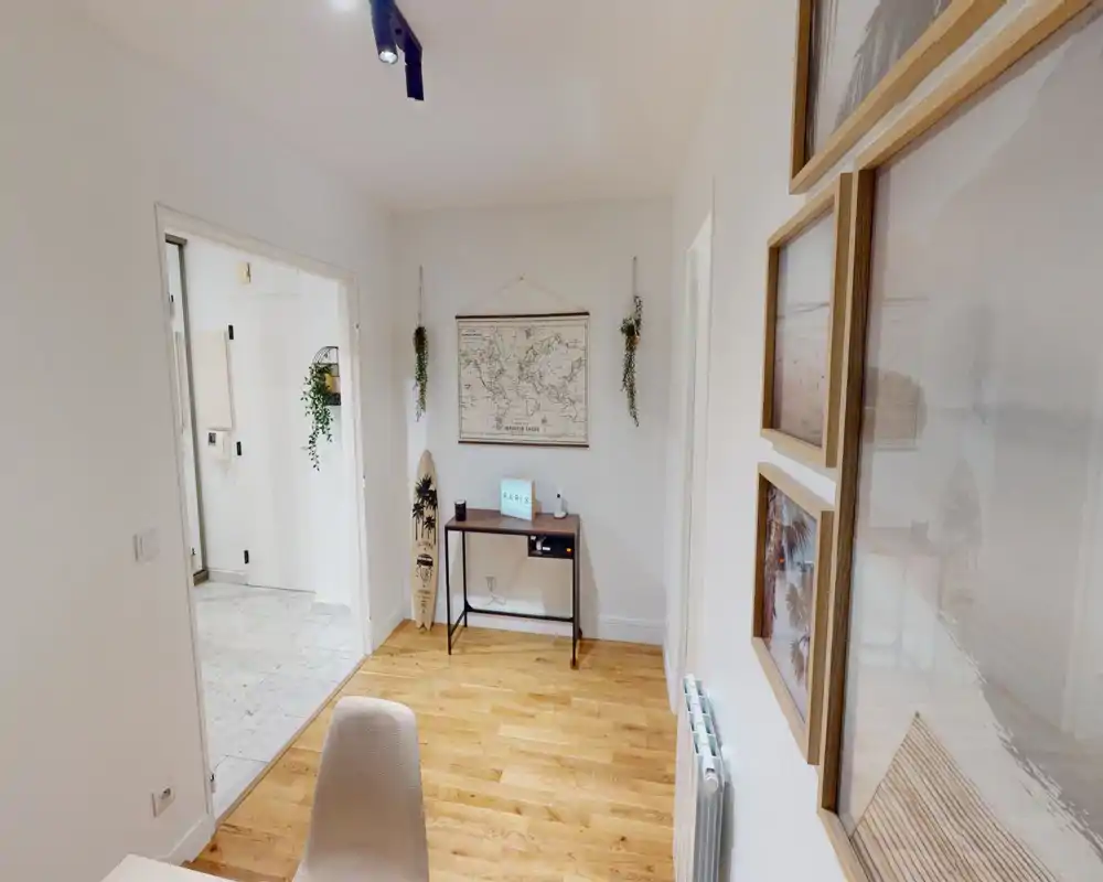 Compact, bright living area with wooden floors, framed wall art, hanging plants and a small console table — a neat, modern corner suitable for relaxation or light work.