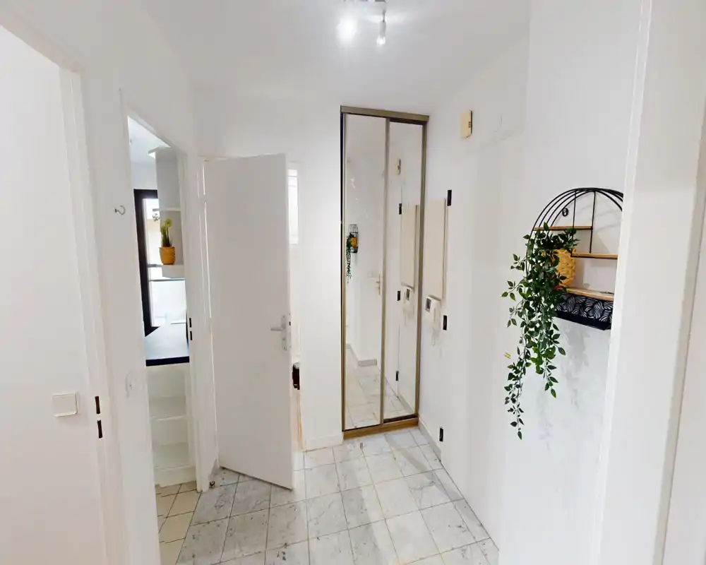 Bright entrance hallway with tiled floor, wall-mounted mirror/closet doors, intercom and decorative shelf with hanging plant — a clean, welcoming entry space.
