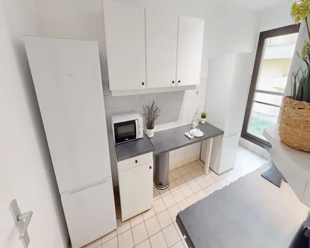 Bright, compact kitchen with white cabinets, two tall refrigerators, a microwave and a clean countertop; large window provides natural light and pleasant plants add a fresh touch.
