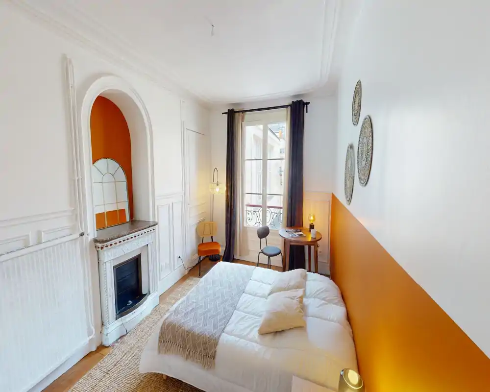 Chambre lumineuse et stylée avec un lit double confortable, un mur orange chaleureux, des assiettes décoratives et une petite table près de la fenêtre — idéale pour un séjour cosy.