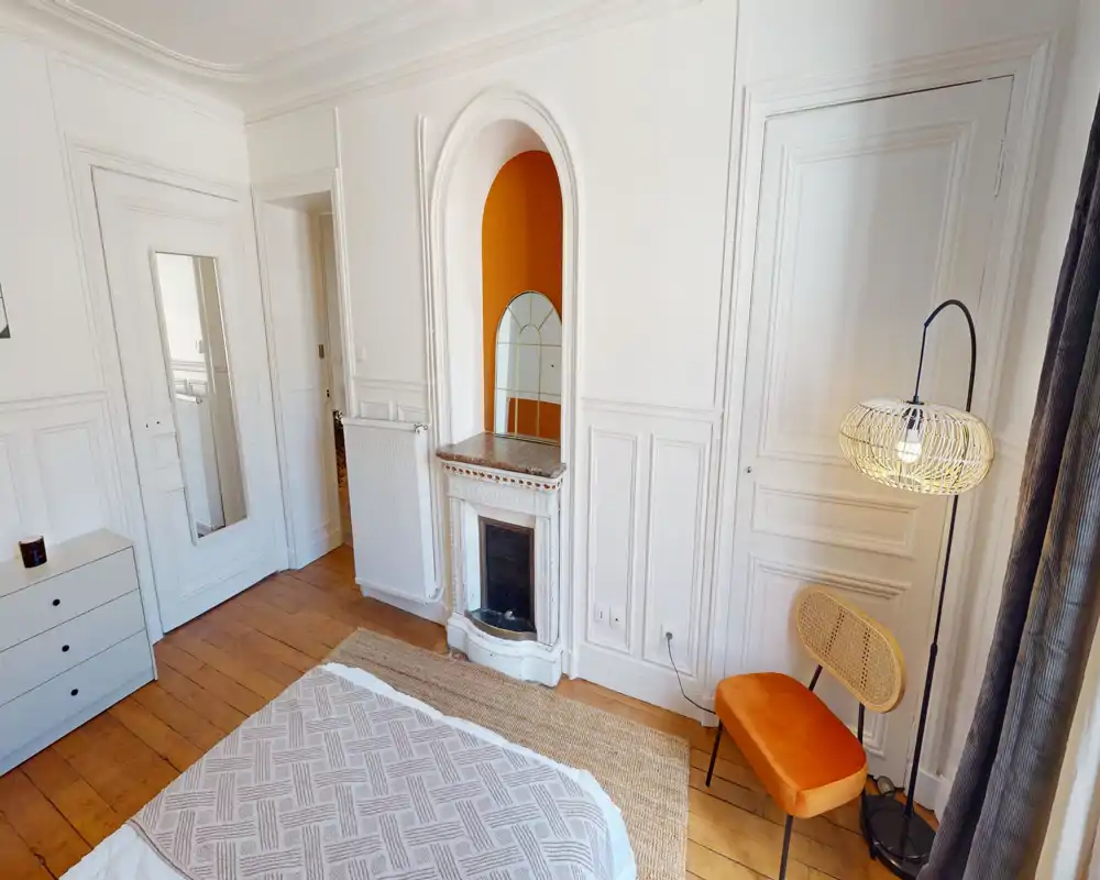 Chambre lumineuse et chaleureuse avec parquet, lit bien fait et accents soignés — chaise en rotin, coussin orange et niche décorative avec cheminée. La lumière naturelle et l’éclairage doux mettent en valeur les moulures élégantes et l’ambiance accueillante.