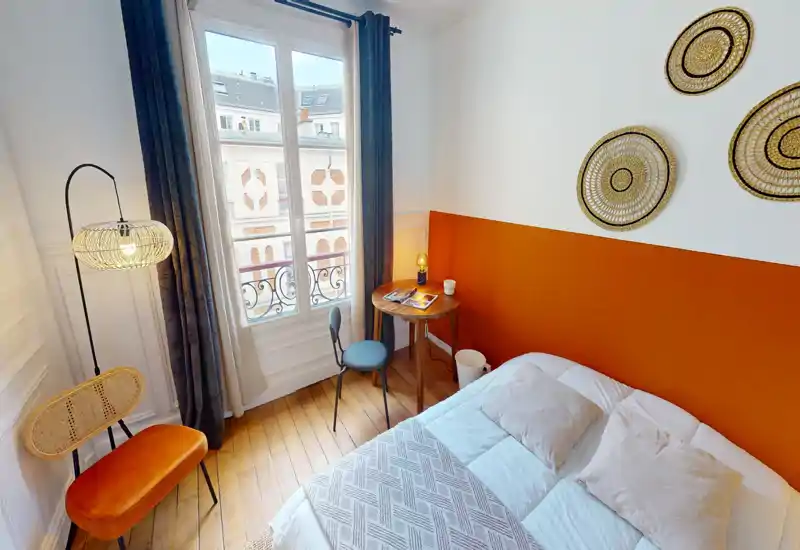 Chambre lumineuse et élégante avec un lit double confortable, un mur d'accent orange et une grande fenêtre apportant beaucoup de lumière naturelle. Textiles douillets, paniers décoratifs et éclairage soigné créent une atmosphère accueillante.