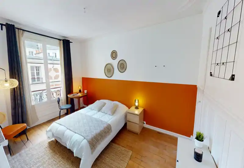 Chambre lumineuse et aménagée avec lit double, mur accent orange et lumière naturelle provenant d'une grande fenêtre. Textiles chaleureux, lampes de chevet et parquet créent une ambiance accueillante parfaite pour une annonce.