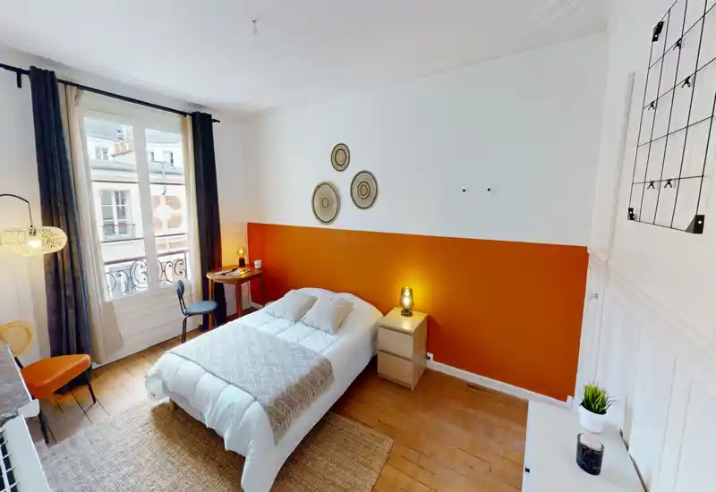 Chambre lumineuse et bien décorée avec un lit double devant un mur orange, lampes de chevet, parquet et grande fenêtre apportant une belle lumière naturelle.