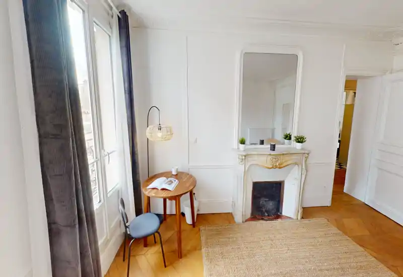 Bright, stylish living area with a decorative fireplace, small round table and seating by a tall window — ideal for relaxing or reading.