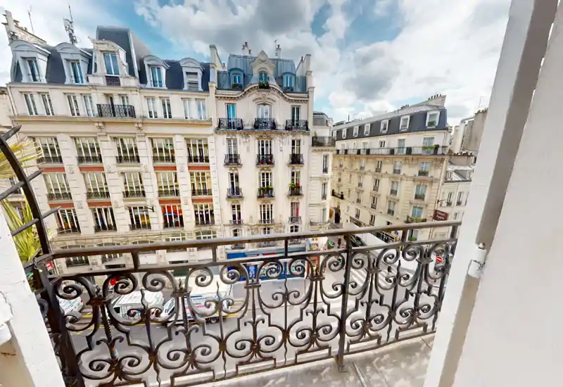 Small Parisian-style balcony with ornate wrought-iron railing overlooking a city street and classic Haussmann buildings, offering a charming urban view.