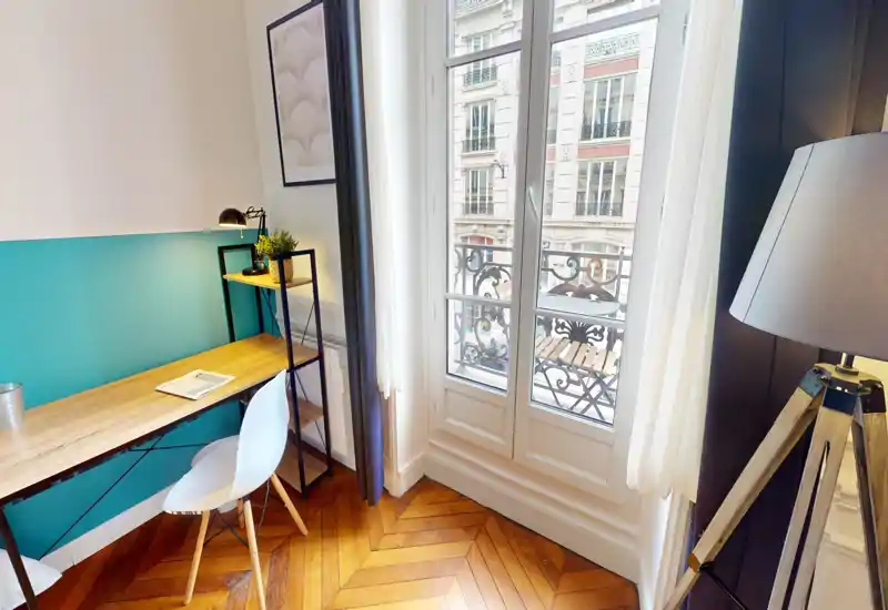 Espace de vie lumineux et élégant avec bureau en bois et chaise près d'une grande fenêtre donnant sur un petit balcon ; décoration moderne, plante et lampadaire trépied pour un coin travail chaleureux.
