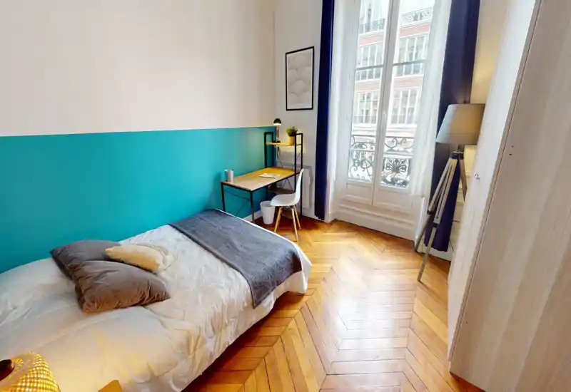 Chambre lumineuse et élégante avec un lit simple confortable, parquet en chevrons et un bureau près de la fenêtre offrant un coin travail agréable.