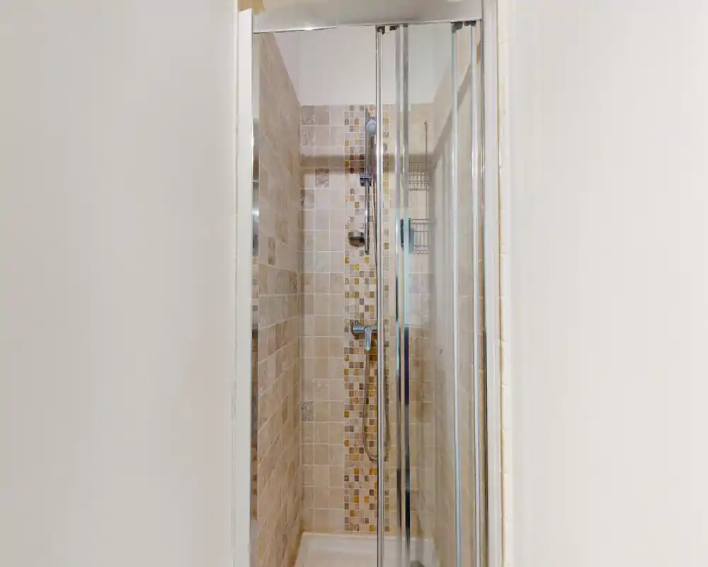 Douche carrelée compacte avec portes coulissantes en verre, robinetterie chromée et bande décorative en mosaïque — salle de bains privée propre et lumineuse.