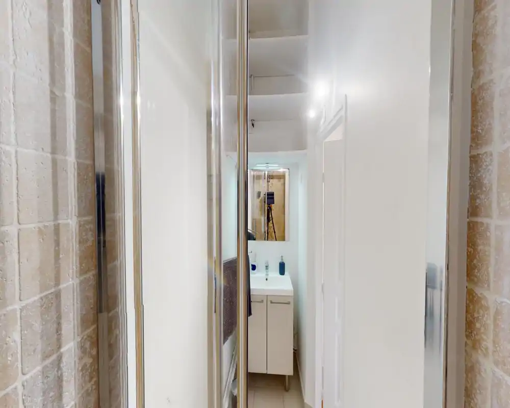 Salle de bains compacte et bien éclairée avec une petite vasque et un miroir ; l'espace douche carrelé est visible à gauche.