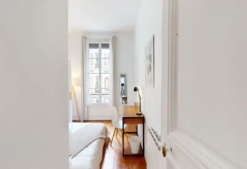 Chambre lumineuse et minimaliste avec lit bien fait, parquet et un petit bureau près de la fenêtre — idéale pour se reposer ou travailler à distance.