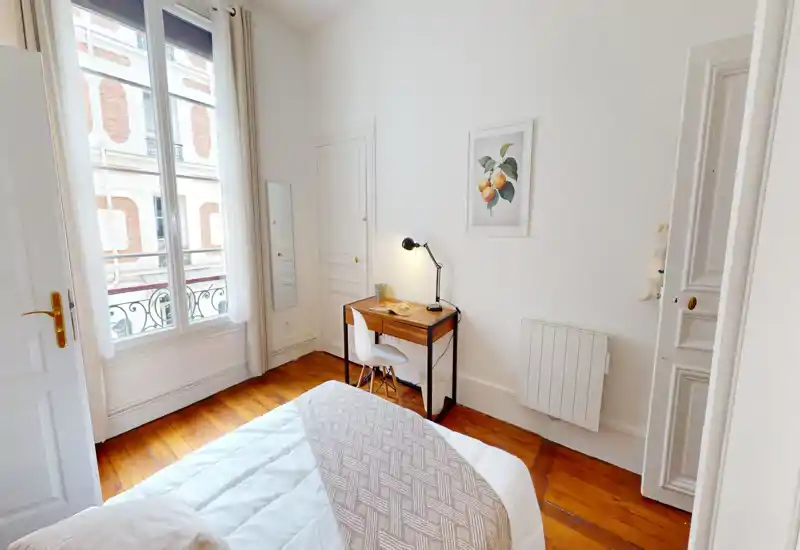 Chambre lumineuse et accueillante avec un lit confortable, parquet et un petit bureau près de la fenêtre. La grande fenêtre apporte beaucoup de lumière naturelle et une décoration minimaliste apaisante.