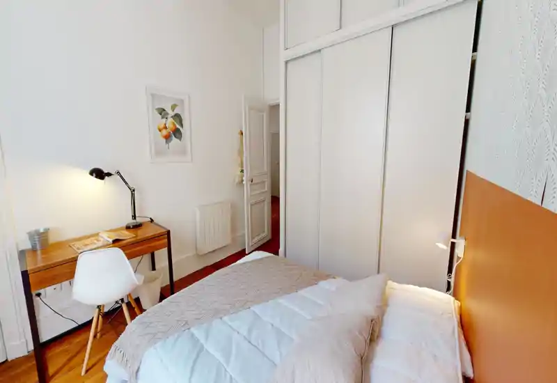Chambre lumineuse et soignée avec un lit double confortable, une tête de lit orange et un petit bureau en bois avec lampe — idéale pour se détendre et travailler.