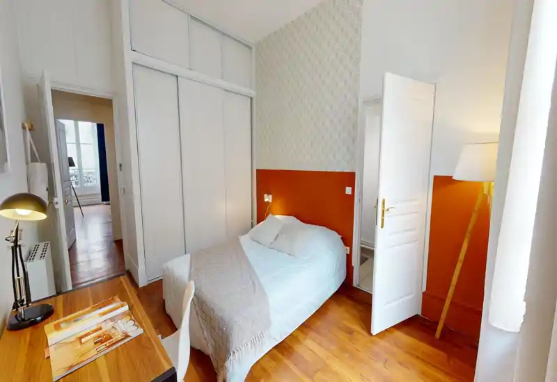 Chambre lumineuse et chaleureuse avec lit double, placard encastré et bureau en bois. Mur accent orange et parquet créent une ambiance accueillante.