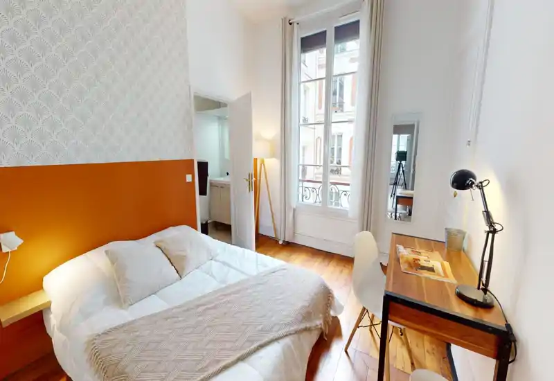 Chambre lumineuse et élégante avec grand lit, mur d'accent chaud et parquet en bois; un bureau avec lampe près de la fenêtre offre un espace de travail pratique.