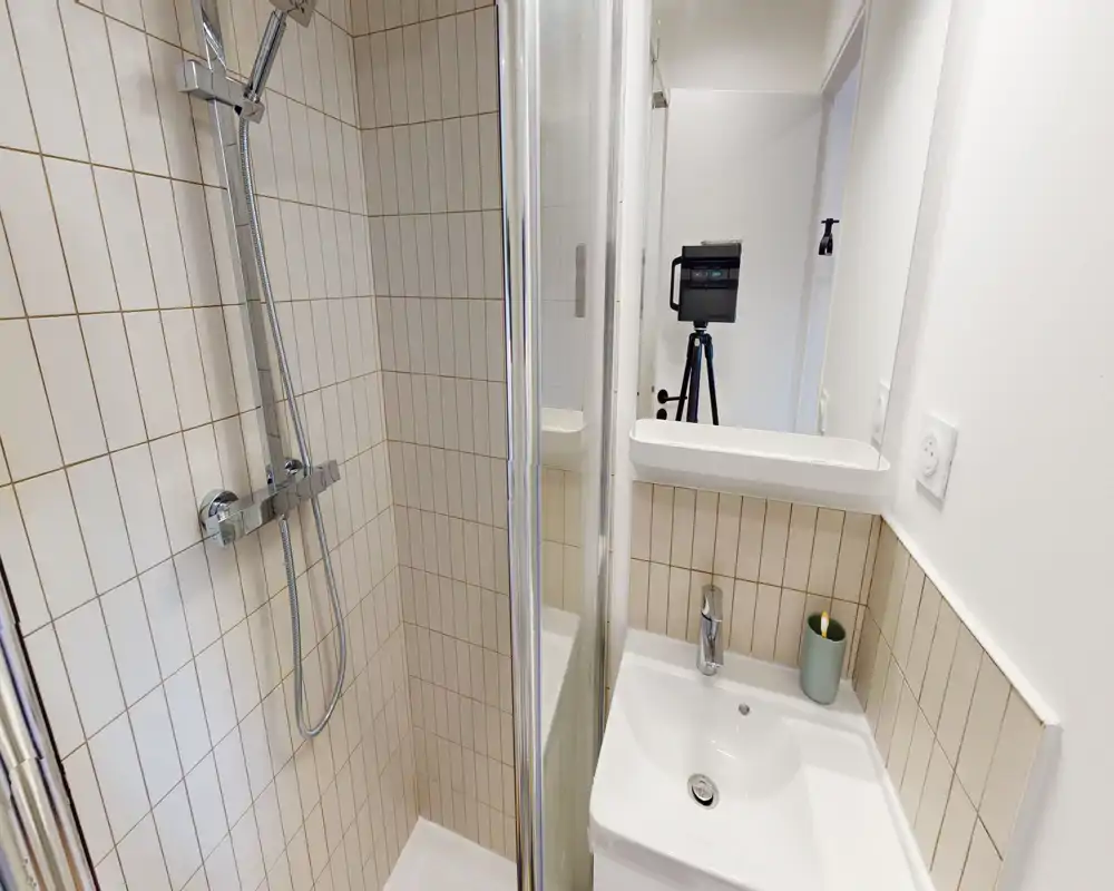 Salle de bains compacte et moderne avec cabine de douche carrelée, douche murale, petit lavabo et miroir. Design propre et lumineux, idéal pour les locations courte durée.