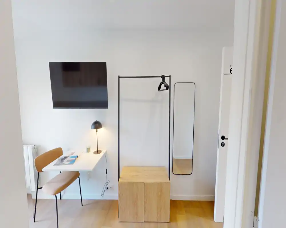 Coin studio lumineux et minimaliste avec TV murale, bureau compact et chaise, portant ouvert avec rangement et miroir pleine longueur — parfait pour une location courte durée moderne.