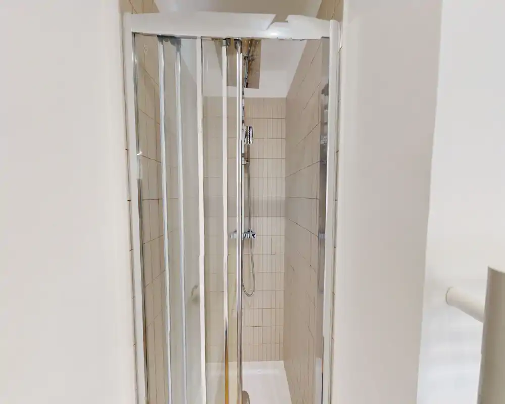 Compact tiled shower with sliding glass doors and a chrome hand shower — clean, modern and well-lit bathroom stall.