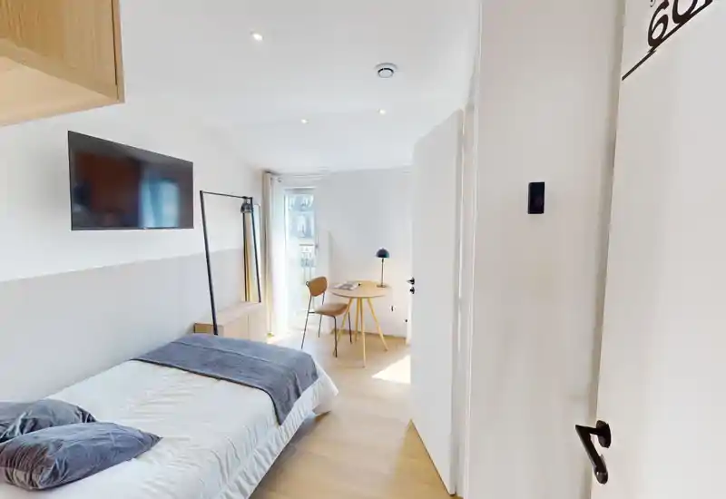Camera da letto moderna e luminosa con un letto singolo ben rifatto, TV a parete e un piccolo angolo studio vicino alla finestra. Tessili morbidi e pavimento in legno donano un’atmosfera pulita e accogliente, perfetta per soggiorni brevi.