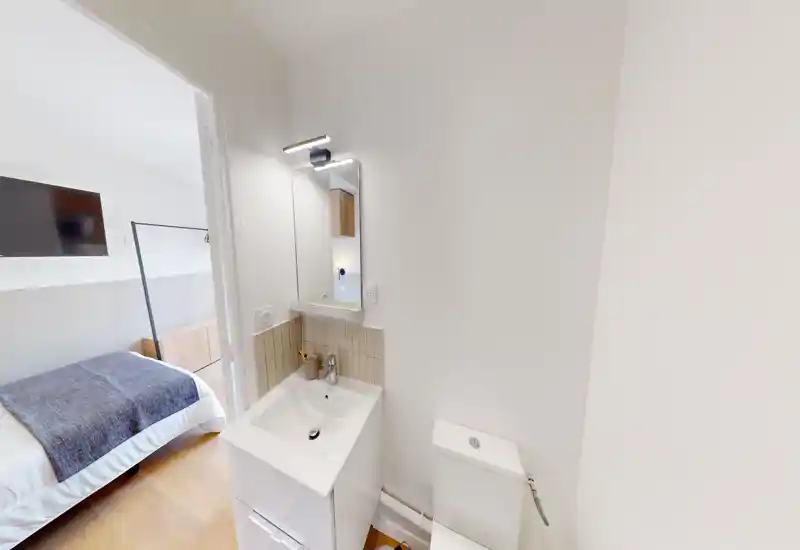 Bagno compatto e moderno con angolo lavabo, specchio e WC; pareti bianche luminose e top piastrellato, porta aperta che conduce ad una camera da letto.