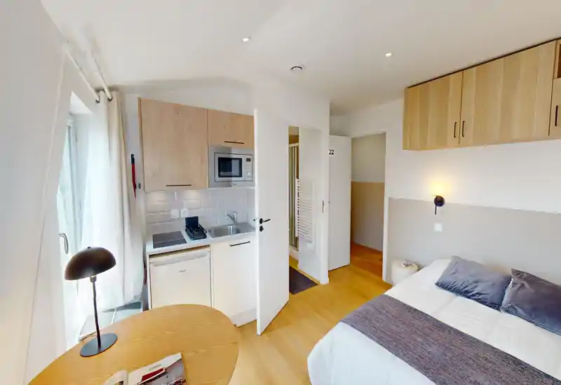 Camera luminosa e compatta con letto matrimoniale, luce da comodino e pensili superiori; presente una piccola angolo cottura e luce naturale dalla finestra — perfetta per un monolocale breve.