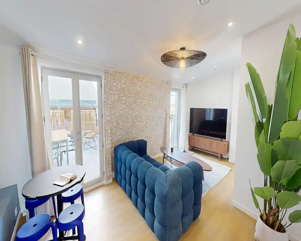 Bright, stylish living room with a deep-blue tufted sofa, wooden coffee table, TV unit and large glass doors opening to a balcony — modern and inviting space ideal for relaxing.