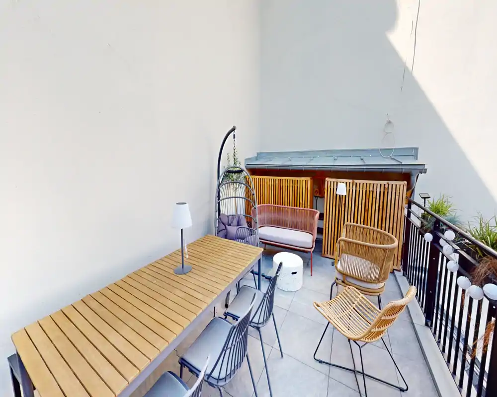 Sunny balcony with a wooden dining table, mixed seating (metal and wicker chairs), a small sofa and decorative plants — ideal for outdoor meals or relaxing.