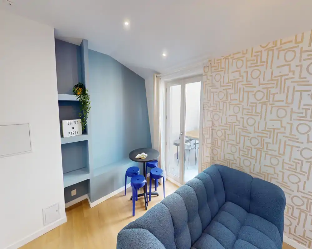Bright, stylish living area with a plush blue sofa, patterned accent wall and a small dining nook next to glass doors leading to an outdoor space.