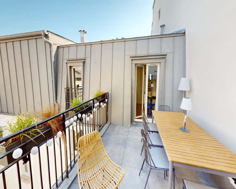 Sunny private balcony with a long dining table, several chairs and a relaxed wicker lounge chair — ideal for alfresco meals or morning coffee.