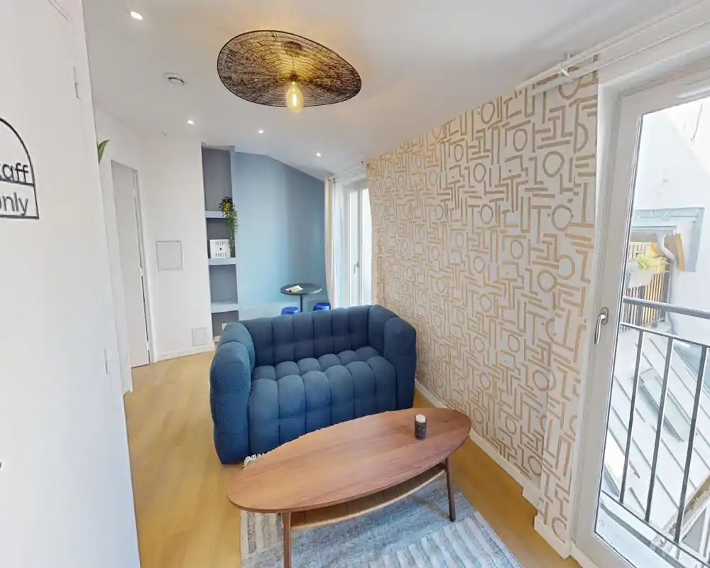 Cozy, stylish small living area with a tufted blue sofa, wooden coffee table and patterned accent wall; bright natural light from the balcony doors enhances the welcoming atmosphere.