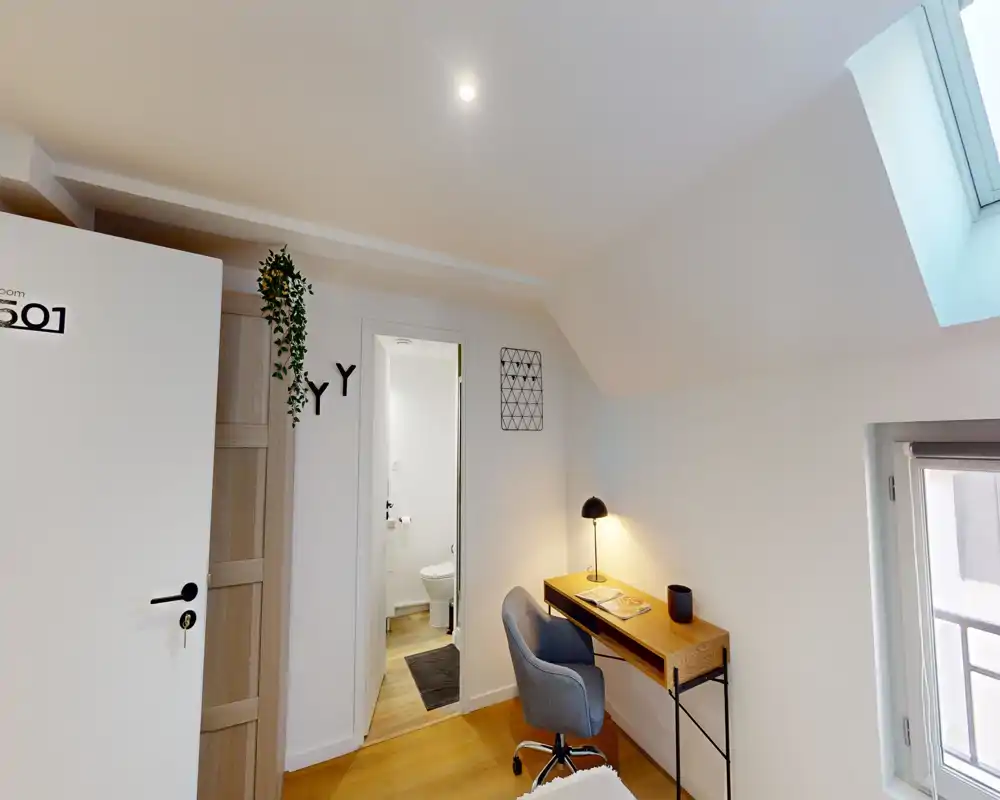 Petit espace mansardé avec bureau en bois, chaise confortable et lampe; puits de lumière et salle de bain attenante visibles par la porte.