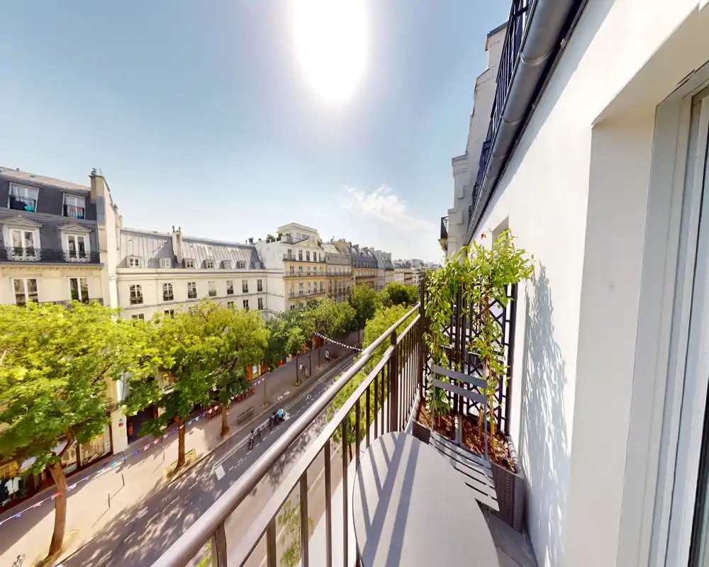 Balcone privato soleggiato con vista su una strada alberata e tipici palazzi parigini; tavolino e vasi rendono l'angolo ideale per un caffè mattutino.