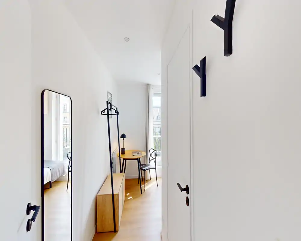 Ingresso luminoso e minimal con appendiabiti a parete, specchio a figura intera, panca bassa e vista parziale sulla camera da letto e un angolo con tavolino rotondo.