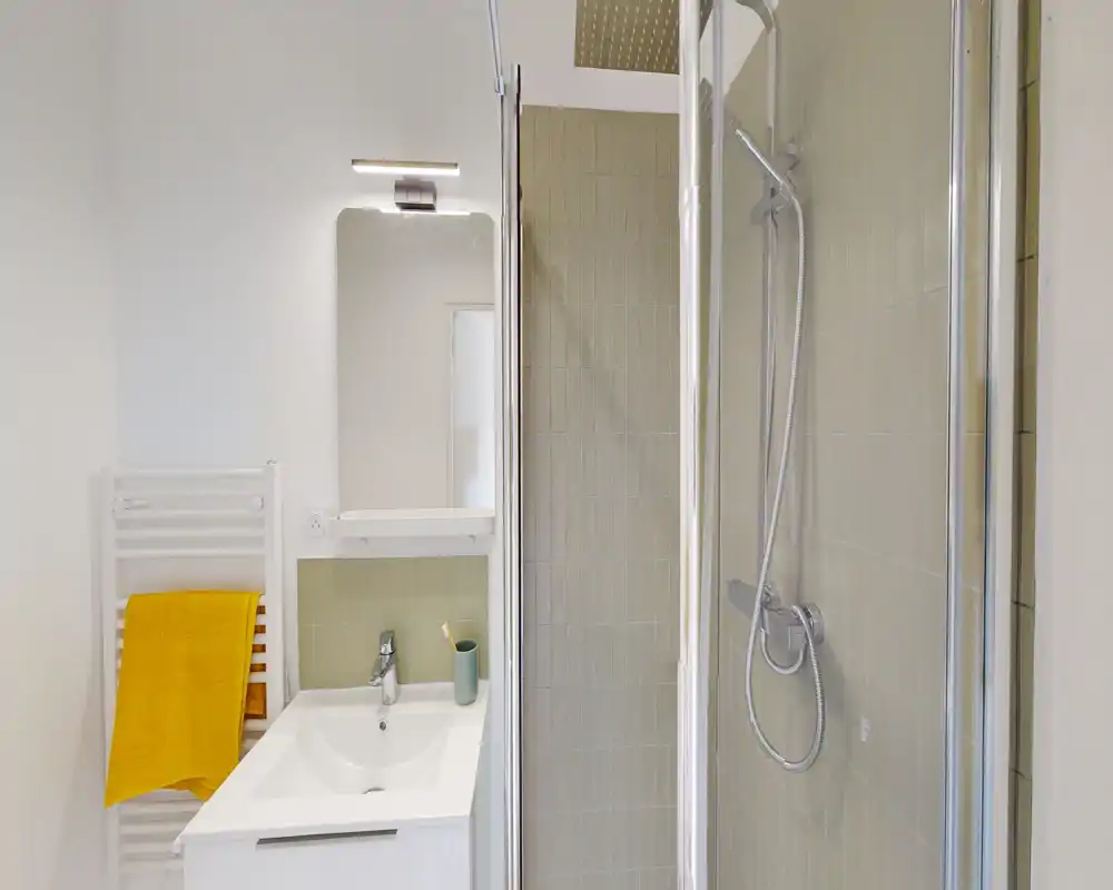 Bagno moderno e luminoso con box doccia in vetro, soffione a parete, lavabo con specchio e scaldasalviette. Piastrelle neutre e un asciugamano giallo come tocco di colore.