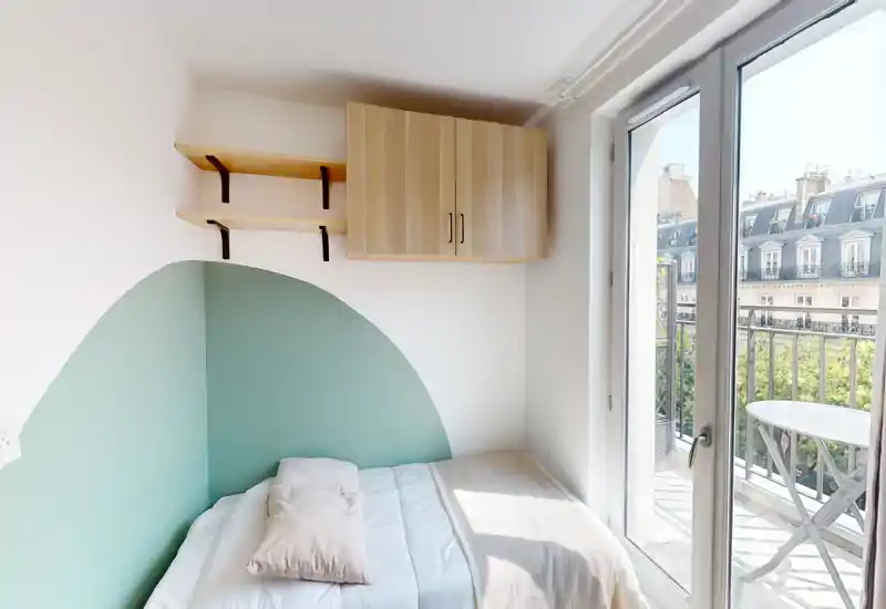 Dormitorio pequeño y luminoso con cama doble, ropa de cama suave y pared con pintura decorativa. Puerta de cristal amplia que abre a un balcón, aportando mucha luz natural y vistas a la ciudad.